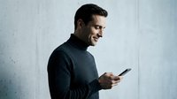 Young adult male, Caucasian, wearing dark charcoal sweater, smiling while holding smartphone against white textured wall background, casual tech lifestyle portrait.