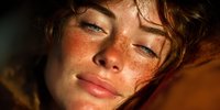 Close-up portrait of young woman with freckles and auburn hair in warm golden sunlight, showcasing natural beauty and glowing skin.