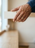 Senior man with weathered hands holding blank white card, wearing blue sleeve and gold wedding band, ideal for business cards, invitations, announcements, and promotional materials.