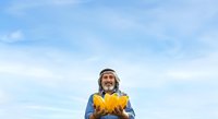 Middle-aged man with long hair and beard, wearing blue shirt and yellow jacket, holding autumn leaves against blue sky background.