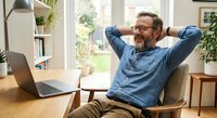 Relaxed mature Caucasian businessman with beard and glasses stretching at desk in bright home office with laptop and plants.