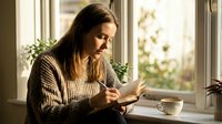 Young woman reading book by window with warm sunlight, cozy sweater, coffee cup on windowsill, peaceful morning routine, relaxation and leisure time.