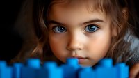 Close up portrait of young girl with bright blue eyes peeking over blue toy blocks against dark background, creating mysterious and innocent atmosphere.