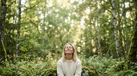 Young woman with blonde hair wearing a cream-colored sweater sits peacefully in a lush forest tunnel, surrounded by green foliage and dappled sunlight, conveying tranquility and connection with nature.