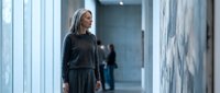 Professional mature blonde businesswoman in dark suit standing confidently in modern corporate office hallway with glass walls and contemporary architecture.