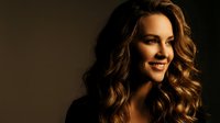 Professional portrait of confident young Caucasian woman with wavy brown hair and warm smile against dark background for business and lifestyle content.