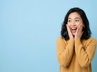 Excited young East Asian woman with dark shoulder-length hair wearing mustard yellow ribbed sweater, hands on cheeks, expressing surprise and joy against blue background for advertising and lifestyle content.