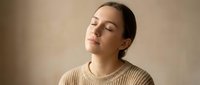 Young woman with closed eyes in peaceful meditation pose wearing beige knit sweater against neutral background for wellness and mindfulness concepts.