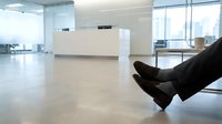 Business professional relaxing in modern office lobby with crossed legs and dress shoes, contemporary reception desk and large windows in background.