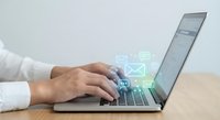 Professional businessperson typing on laptop with digital communication icons floating above keyboard showing email, messaging and connectivity symbols for modern workplace.