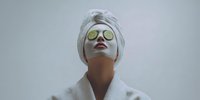 Young woman in spa bathrobe and head towel wearing cucumber eye mask during beauty treatment against neutral background.