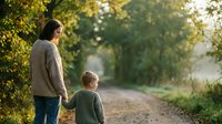Father and son walking together on rural dirt road surrounded by lush green trees during golden hour sunset creating warm family bonding moment in countryside.