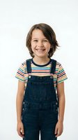 Happy young child with brown hair wearing denim overalls and striped shirt smiling at camera on white background isolated for design use.