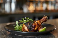 Grilled chicken leg with caramelized glaze served on black plate with fresh microgreens and lime wedge garnish against dark restaurant background.