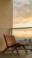 Modern wooden lounge chair on high-rise balcony overlooking cityscape at sunset. Minimalist interior design with floor-to-ceiling windows framing golden sky and urban skyline for contemporary home decor.