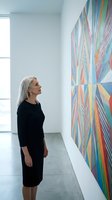 Mature Caucasian woman in black dress admiring colorful abstract artwork in modern gallery space with natural light from window.