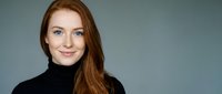 Young professional Caucasian woman with long red hair wearing black turtleneck sweater smiling confidently at camera against neutral gray background.