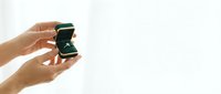 Young adult with medium skin tone holding open a green velvet ring box containing a diamond engagement ring against white background, suitable for wedding, proposal, and jewelry marketing.