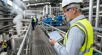 Senior industrial engineer in safety vest and hard hat reviewing technical documentation on tablet while overseeing manufacturing plant operations and equipment.