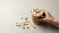 Hot chocolate with marshmallows in ceramic mug held by hand on beige background. Cozy winter drink with wooden spoon and scattered marshmallows.
