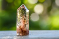 Natural rutilated quartz crystal point with copper-colored inclusions stands on wooden surface against blurred green bokeh background, displaying mystical properties.