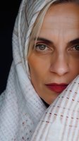 Close-up portrait of young woman with green eyes and red lipstick wearing white patterned headscarf, looking directly at camera with intense gaze against dark background.