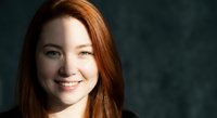 Young redhead woman with long hair smiling warmly at camera against dark textured background for portrait photography and lifestyle content.