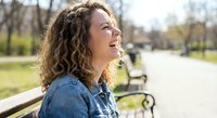 Happy young woman with curly hair laughing outdoors in park wearing denim jacket on sunny day expressing joy and positive emotions for lifestyle content.