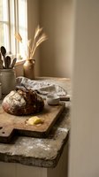Artisanal sourdough loaf with crispy crust on wooden cutting board in warm sunlit kitchen with dried wheat arrangement and baking tools for bakery and food styling projects.