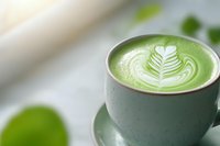Green matcha latte with white foam art leaf pattern in sage ceramic cup on light background, selective focus creates soft dreamy atmosphere.