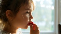 Young girl with light brown hair and fair complexion, appearing anxious or worried, biting her red nail while gazing out window, suitable for mental health, stress, and childhood emotion content.