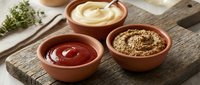 Three terracotta bowls filled with condiments including ketchup, mustard and seasoning blend arranged on rustic wooden cutting board with herbs for cooking.