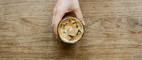 Person's hand holding creamy peanut butter in glass jar on wooden surface, overhead view for food styling, cooking, and recipe content.