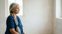 Senior woman with gray hair in braids, wearing blue cardigan, standing in bright interior space with natural light from windows, contemplative expression, suitable for healthcare, wellness, aging, lifestyle and editorial content.