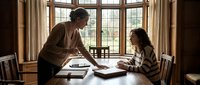 Young Caucasian female tutor teaching Asian student girl at wooden table near large windows in warm library setting for educational content.
