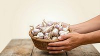 Hands holding wicker basket filled with fresh garlic bulbs on wooden surface against neutral background. Suitable for cooking, agriculture, and food-related designs.