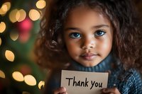 Young girl with striking blue eyes and curly hair holding thank you note card against blurred Christmas lights background creating warm bokeh effect.