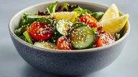 Fresh vegetable salad with cherry tomatoes, cucumber slices, and arugula leaves garnished with sesame seeds and lemon wedges in gray ceramic bowl.