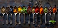 Aromatic spices and herbs in vintage metal spoons on dark rustic background, colorful seasoning collection arranged in line for cooking and food styling.
