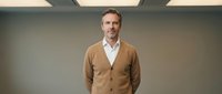 Professional middle-aged Caucasian businessman in brown cardigan and white shirt standing confidently in modern office environment for corporate presentations.