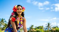 Young woman with South Asian ethnicity wearing vibrant floral lei and tropical flower crown, smiling outdoors against blue sky and palm trees. Summer vacation and Hawaiian culture concept for travel and lifestyle.