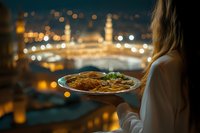 Young woman holding plate with traditional Middle Eastern food on balcony overlooking evening cityscape with glowing lights and mosque silhouette in background.
