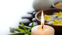 Spa wellness scene with burning candle, hot stones, flower petals in water bowl and bamboo leaves for relaxation and massage therapy treatments.