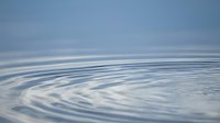 Gentle water ripples create concentric circles on calm blue surface reflecting cloudy sky. Peaceful aquatic background for meditation and wellness concepts.