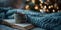 Steaming cup of coffee on open book with cozy blue knitted blanket against blurred Christmas lights background, creating warm winter atmosphere.