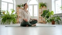 Young Caucasian woman practicing meditation in lotus pose on yoga mat in bright room with houseplants, mindfulness and wellness lifestyle.