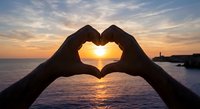 Hands forming heart shape silhouette against golden sunset over ocean water with lighthouse in background creating romantic peaceful moment.