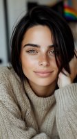Young brunette woman in cozy beige sweater smiling warmly at camera with natural makeup and tousled hair in comfortable indoor setting.