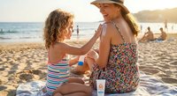 Mother and daughter applying sunscreen on sunny beach during summer vacation. Woman in hat helping child with sun protection while sitting on blanket by ocean.