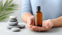 Female hands holding brown glass essential oil bottle against light background with zen stones and palm leaf, wellness and aromatherapy concept for spa treatments.